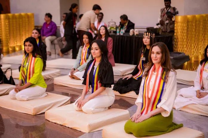 Women in sashes seated on cushions in a hall with an audience behind