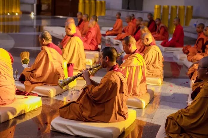 Robed figures seated on the floor of a hall, one playing a wind instrument